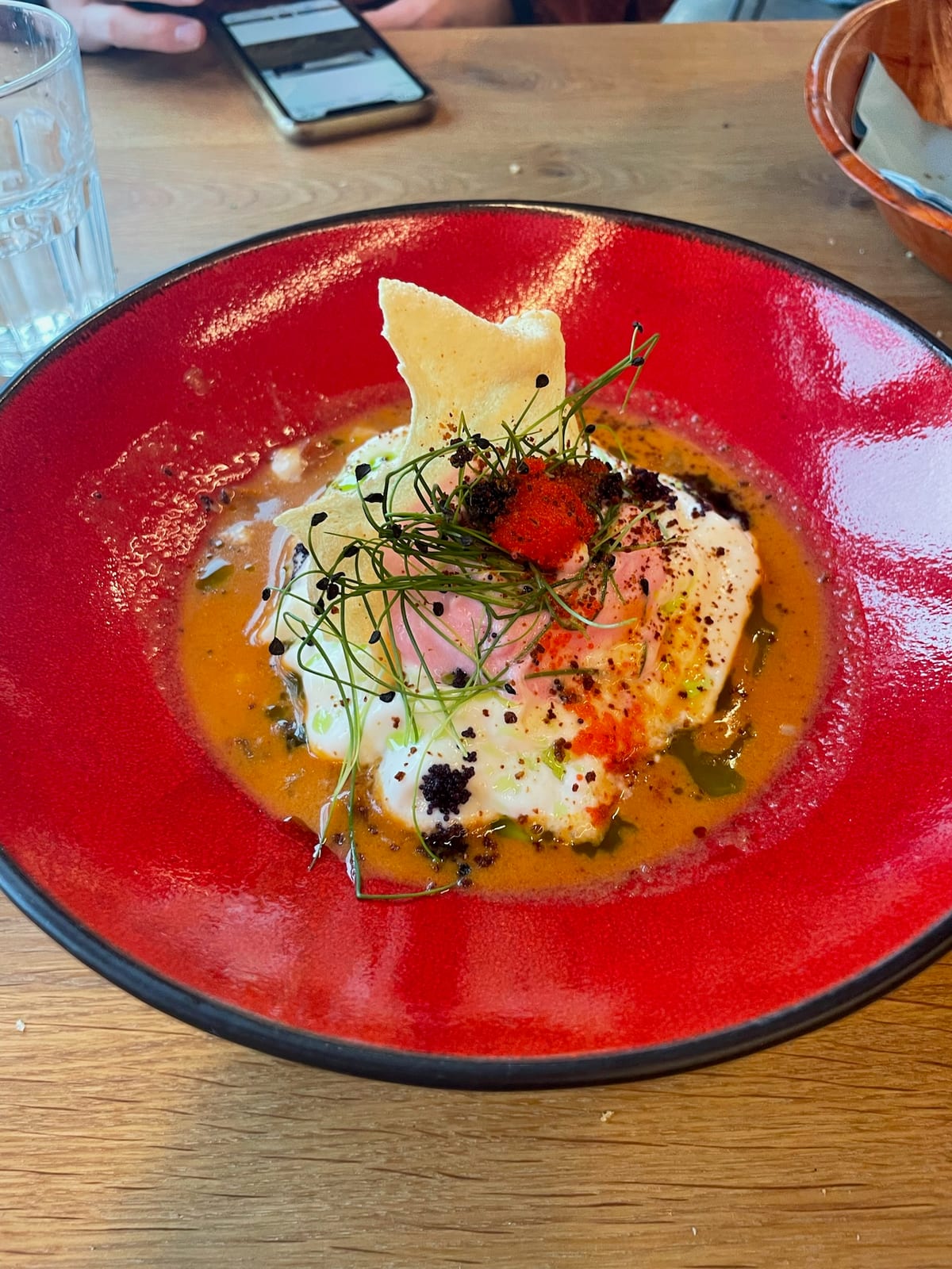 A plated fish dish in a red bowl, dressed with herbs, roe, and a crisp